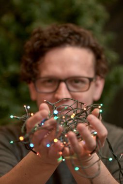 İki elinde renkli Noel ışıkları tutan gözlüklü beyaz bir genç adam. Seçici odak.