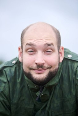 Uykulu bir gülümsemeyle kameraya bakan genç bir adamın portresi. O dışarıda ve ceketi yağmurdan ıslanmış..