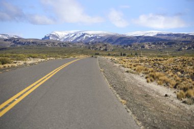 Asfalt karayolu engebeli bir patagonya arazisi boyunca uzanan altın çimenler, kayalık tepeler ve açık mavi gökyüzünün altındaki uzak karla kaplı dağlar, macera ve yolculuğu simgeliyor.