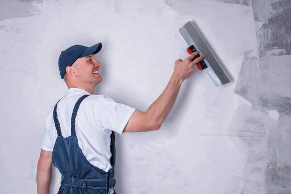 Smiling worker in profile wearing overalls and cap plastering a wall with finishing putty using a putty knife. Repair work and construction concept