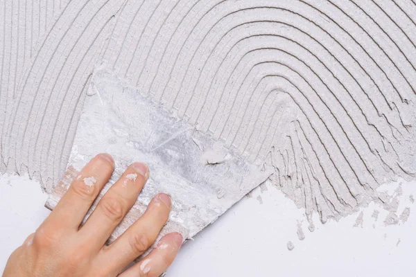 Male hand, soiled with a building mixture, holding a comb putty-knife and covering a wall surface with adhesive paste before laying a tile or decorative stone. Repair work and renovation concept