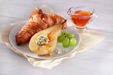 Sabah ışığında beyaz ahşap masada taze kruvasan, üzüm, pişmiş armut mavi peynir ve bal ve bal ile cam tabak ile doldurulmuş beyaz tabak