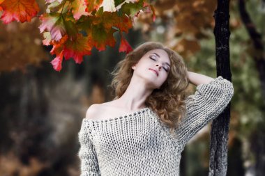 Sonbahar parkında renkli yeşil ve kırmızı akçaağaç ağacının yanına atılmış bir kafa ile örme kazak güzel bir uzun saçlı sarışın kız Portresi. Sonbahar manzarası