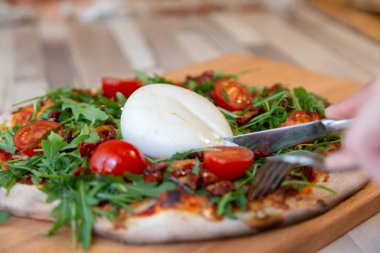 Bir çatal ve bıçakla peynirli bir Pizza domates, roka ve burrata ahşap bir gemide arka plan için ahşap bir masa ve tuğla duvar üzerine peynir kesme holding eller
