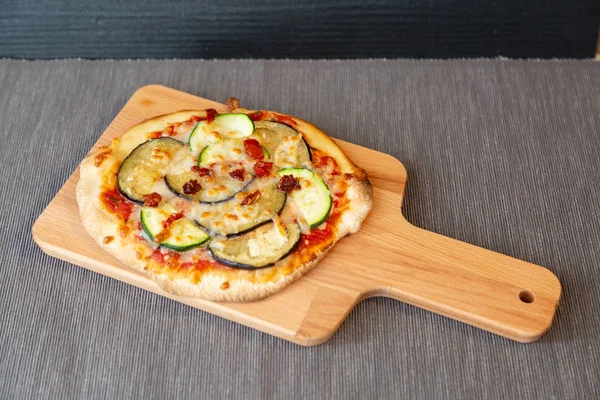 Aerial view of a pizza with zucchini, eggplant and dry tomatoes with gratin mozzarella cheese. On a wooden board and gray tablecloth