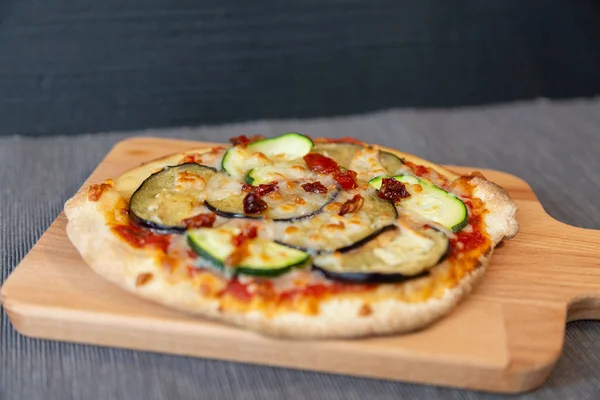 Close up of a pizza with zucchini, eggplant and dry tomatoes with gratin mozzarella cheese. On a wooden board and gray tablecloth