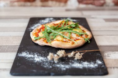 roka, peynir ve domates bir kara tahta üzerinde pizza un ile garnitür ve bir tuğla duvar ile ahşap bir masaya çiçek Kuru.