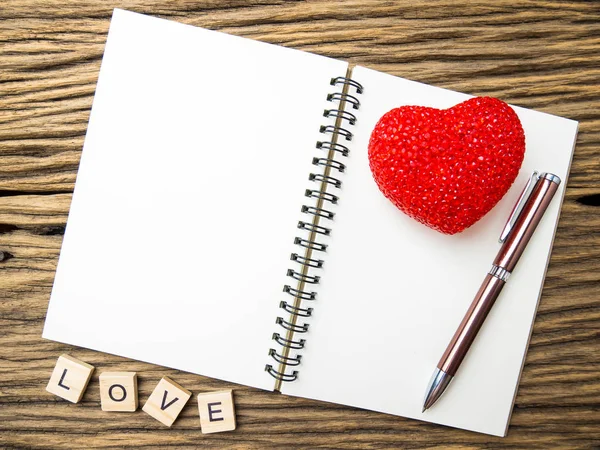 Top view of notebook and pen with red heart on wooden blackground, LOVE word in wooden cube, Valentine\'s Day.