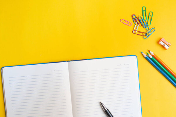 Notepad with pen and pencils on yellow background. Colorful paper clips. Back to school concept. Copyspace.
