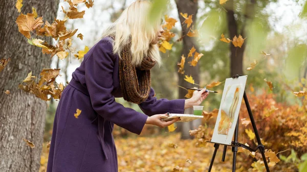Blonde girl artist draws among falling leaves in the autumn park