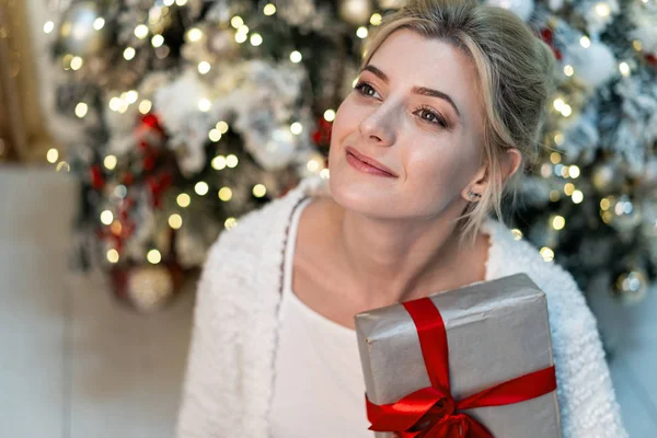 Half length portrait of beautiful young blonde girl in white sweater posing with gift near the Christmas tree