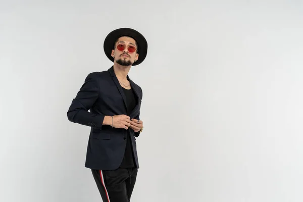 Retrato de mitad de longitud de un joven guapo y moderno hombre vestido con traje negro y sombrero en gafas de sol redondas — Foto de Stock