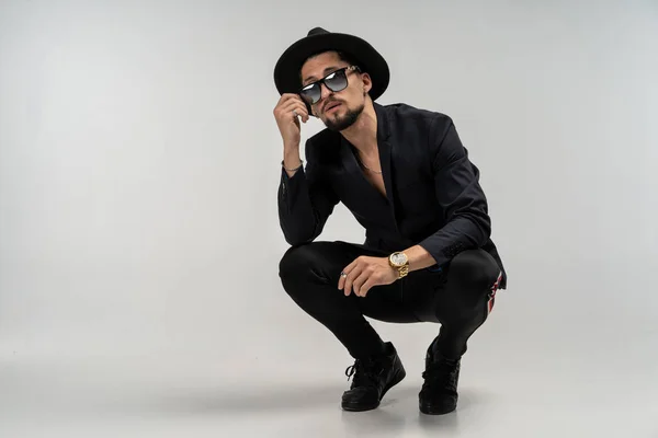 Stylish man in black suit and hat in round sunglasses crouched and posing