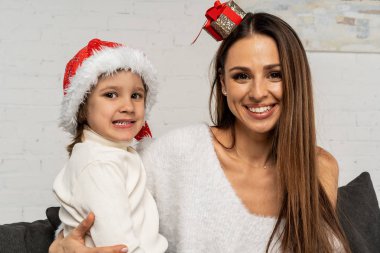 Küçük oğlu santa şapka sarılma beyaz kazaklı genç gülümseyen anne portresi yarım uzunluk