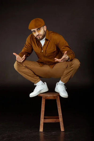 Stylish young man in beige clothes and yellowe sunglasses standing on chair looking at the camera, posing on black background