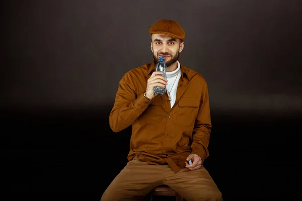 Casualmente guapo hombre en sombrero beige sentado en la silla y agua potable — Foto de Stock