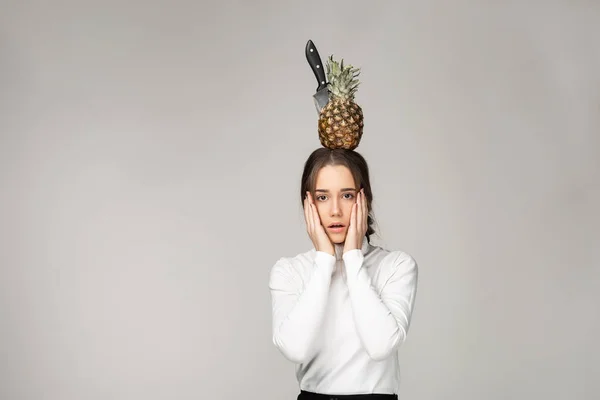 Surprised young girl holds cheeks by hands, pineappple with knife on her had