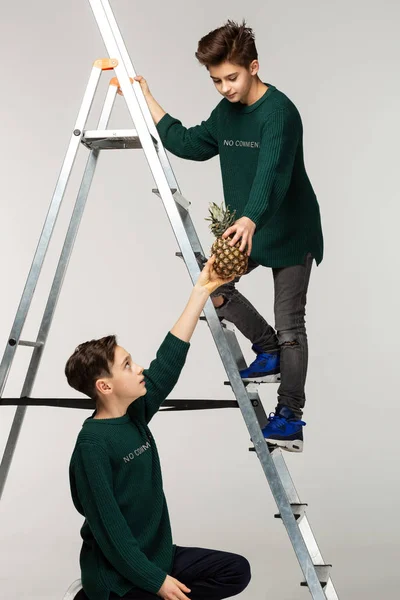 Two boys teenagers in green sweaters, one is standing on ladder other gives him pineapple