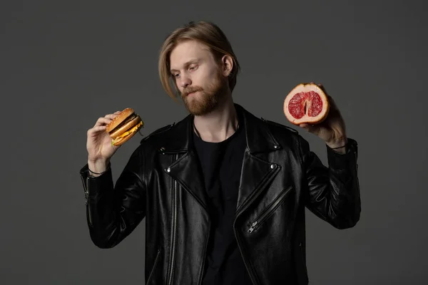 Aantrekkelijke bebaarde jonge man in leren jas kiest tussen een hamburger en grapefruit — Stockfoto
