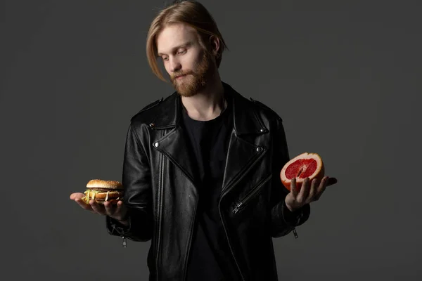 Atractivo joven barbudo en chaqueta de cuero elige entre una hamburguesa y pomelo — Foto de Stock