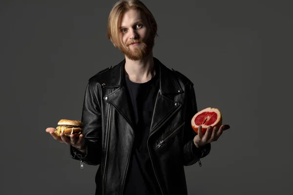 Aantrekkelijke bebaarde jonge man in leren jas kiest tussen een hamburger en grapefruit — Stockfoto