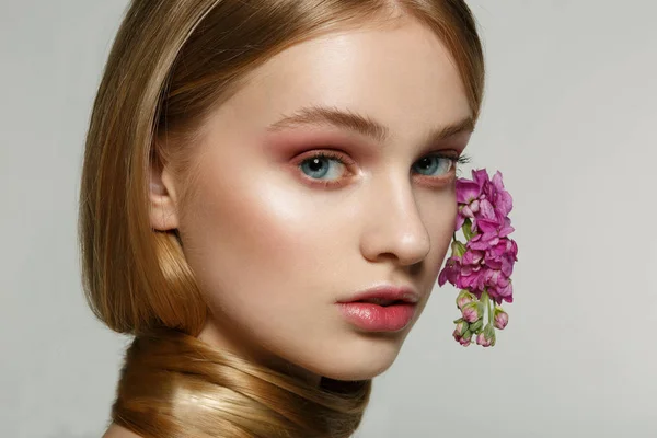 Gros plan portrait de jeune fille aux yeux bleus, maquillage lumineux, cou enveloppé dans les cheveux, fleurs violettes frisées dans les cheveux — Photo