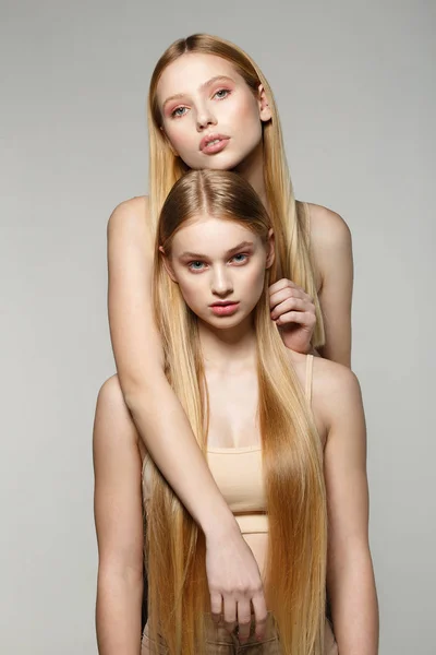 Dvě krásné blondýnky s dlouhými vlasy na kameru v ateliéru při pohledu na světlé pozadí — Stock fotografie