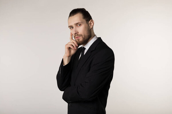 Side view portrait of attractive young businessman in black suit with stylish hairstyle holding hand on chin with thoughtful expression