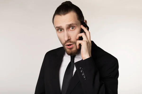 Retrato de cintura hacia arriba de un joven hombre de negocios barbudo en traje negro sorprendido por las noticias escuchadas de un socio de negocios — Foto de Stock