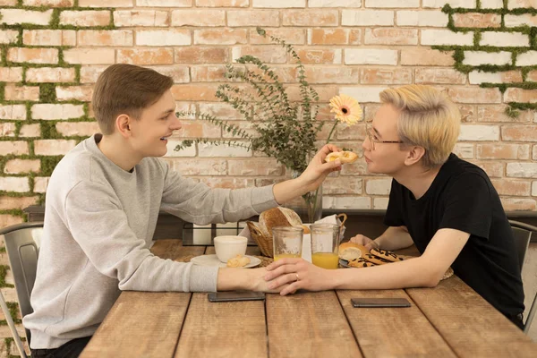 Το ευτυχισμένο ζευγάρι έχει πρωινό στην κουζίνα του σπιτιού. Όμορφοι ομοφυλόφιλοι άνδρες πίνοντας χυμό και ταΐζοντας ο ένας τον άλλον — Φωτογραφία Αρχείου