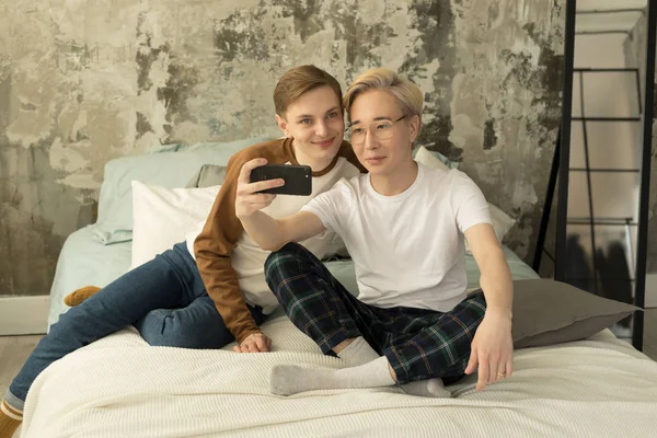 Atraktivní mezinárodní homosexuální pár relaxační v posteli před nočním spánkem a pohled na obrazovku smartphone — Stock fotografie