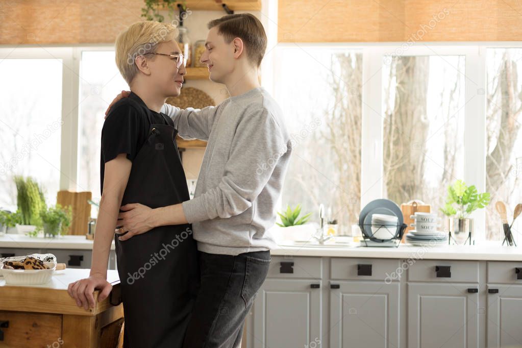 International couple of two young mens in casual clothes hugging in the kitchen