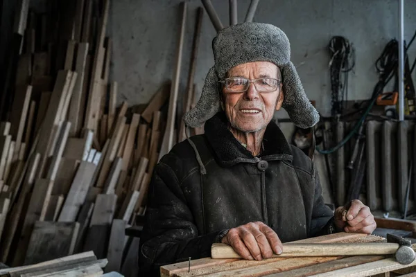 Hombre muy viejo maestro en ropa de abrigo gris y gafas con martillo en las manos —  Fotos de Stock