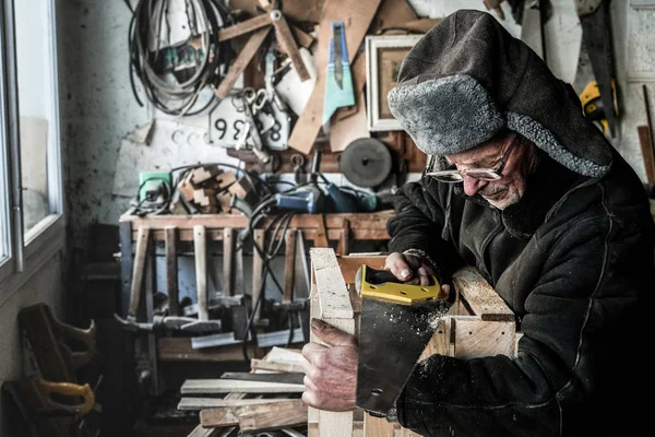 Ritratto in vita del nonno anziano in abiti grigi caldi in occhiali segare tavola di legno — Foto Stock