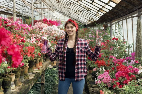 Mujer morena feliz jardinero con flores rosas y rojas en un invernadero con camisa a cuadros con una diadema roja — Foto de Stock