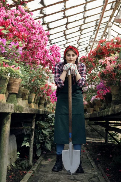 Niña jardinero en delantal y guantes con una gran pala en invernadero, mirando a la cámara — Foto de Stock