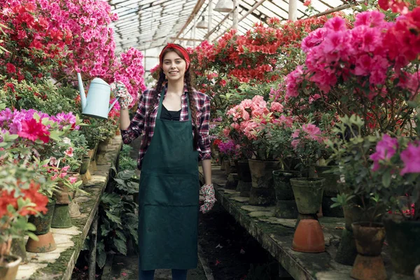 Atractiva joven jardinero en ropa de trabajo con diadema roja regando flores de colores en invernadero — Foto de Stock