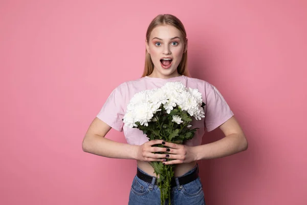 Jeune femme blonde surprise à bouche ouverte portant un t-shirt rose avec des fleurs blanches dans les mains — Photo