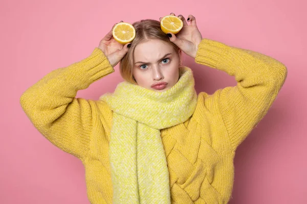 Человеческие эмоции. Блондинка красивая европейская девушка в желтом свитере и шарфе с забавным лицом, держащая на голове два нарезанных цитрусовых лимона — стоковое фото