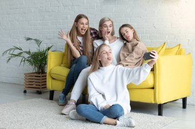 Evde sarı kanepede otururken selfie yaparken rahat giysiler içinde Mutlu çekici lezbiyen aile, sarışın daughtet çapraz bacaklar ile katta oturan ve elinde telefon tutarak