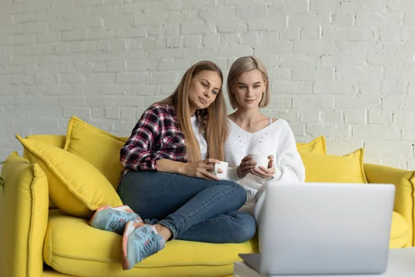 Två vackra lesbisk kvinna i casual kläder sitter tillsammans på gula soffan hemma — Stockfoto