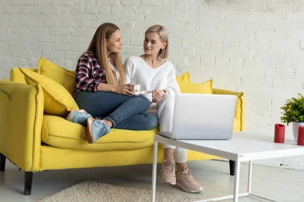 Härlig lesbisk par i casual kläder sitter tillsammans på gul soffa med kopp te — Stockfoto