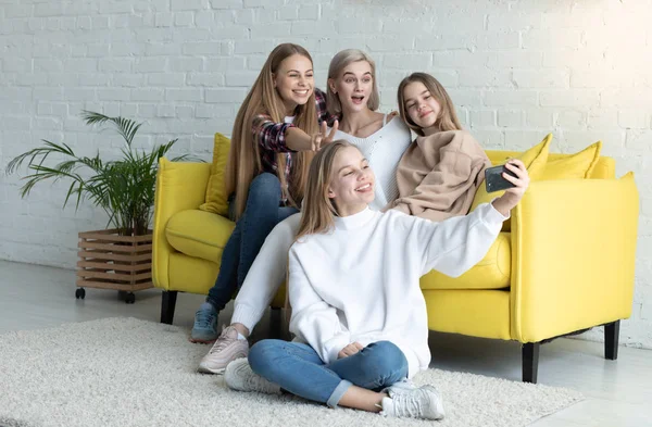 Lycklig attraktiv lesbisk familj i casual kläder gör Selfie medan du sitter på gul soffa hemma, Blonda daughtet sitter på golvet med korsade ben och hålla telefonen i handen — Stockfoto