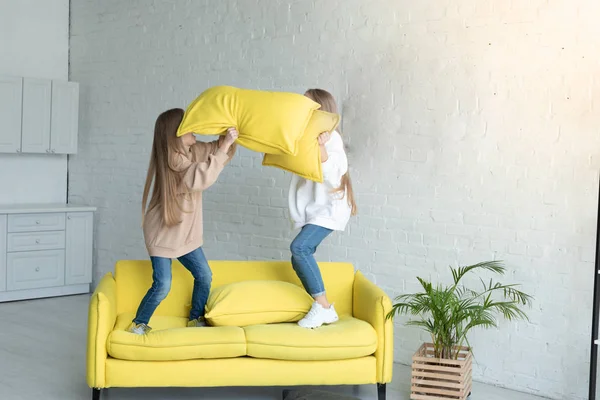 Två unga systrar i casual kläder gjorde kudde slagsmål, hoppa tillsammans på soffan — Stockfoto