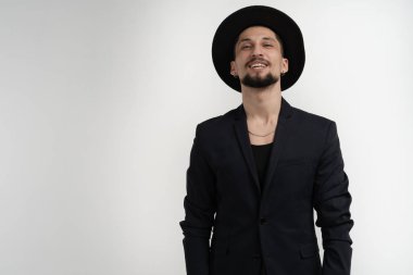 Sakallı mutlu neşeli genç adam, siyah trendy takım elbise ve şapka gülümseyerek giyen ve beyaz arka plan üzerinde izole kamera bakarak