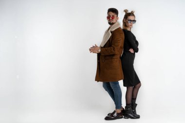 Sırt sırta durarak beyaz arka planda izole edilmiş kameraya bakarak şık ve modaya uygun giyinen genç bir çift.