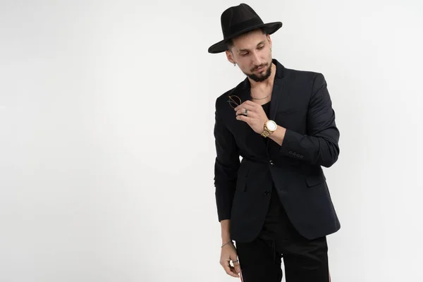 Retrato de moda de hombre elegante guapo en traje negro y sombrero con gafas de sol en la mano, mirando hacia abajo, posando en estudio sobre fondo gris — Foto de Stock