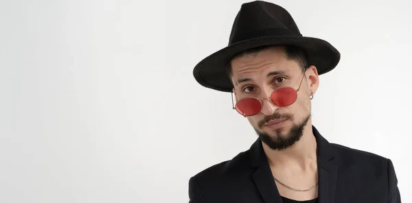 Joven barbudo alegre confiado en traje negro y sombrero en gafas de sol rojas aisladas sobre fondo blanco — Foto de Stock