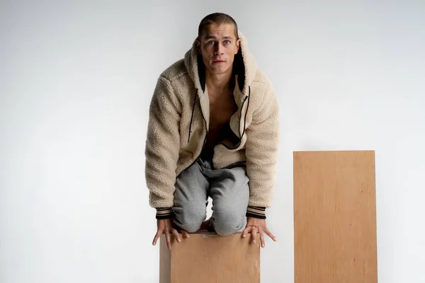 Jovem atraente homem com corte de cabelo curto, vestindo sportswear, ajoelhado em cubo de madeira, olhando para a câmera com rosto engraçado isolado sobre fundo branco — Fotografia de Stock