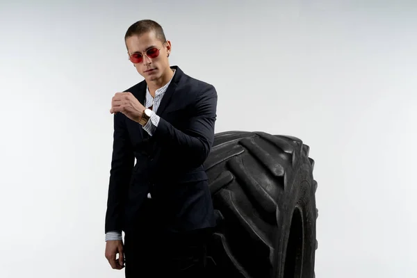 Hombre joven guapo en traje negro y gafas de sol rojas de pie delante de un neumático grande, mirando a la cámara, aislado sobre fondo blanco, espacio de copia para su texto — Foto de Stock